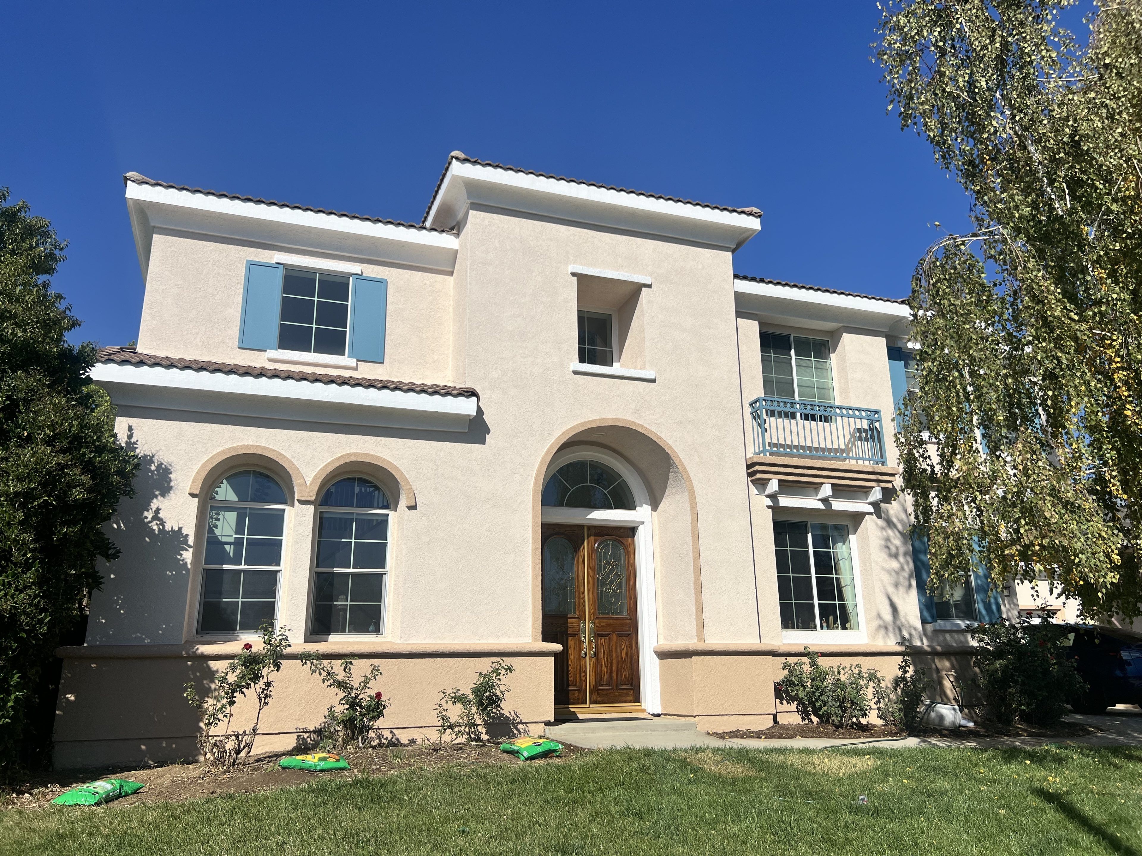 Two-story stucco house with blue shutters, arched windows, and wooden door. Surrounded by green grass and trees, under a clear blue sky.