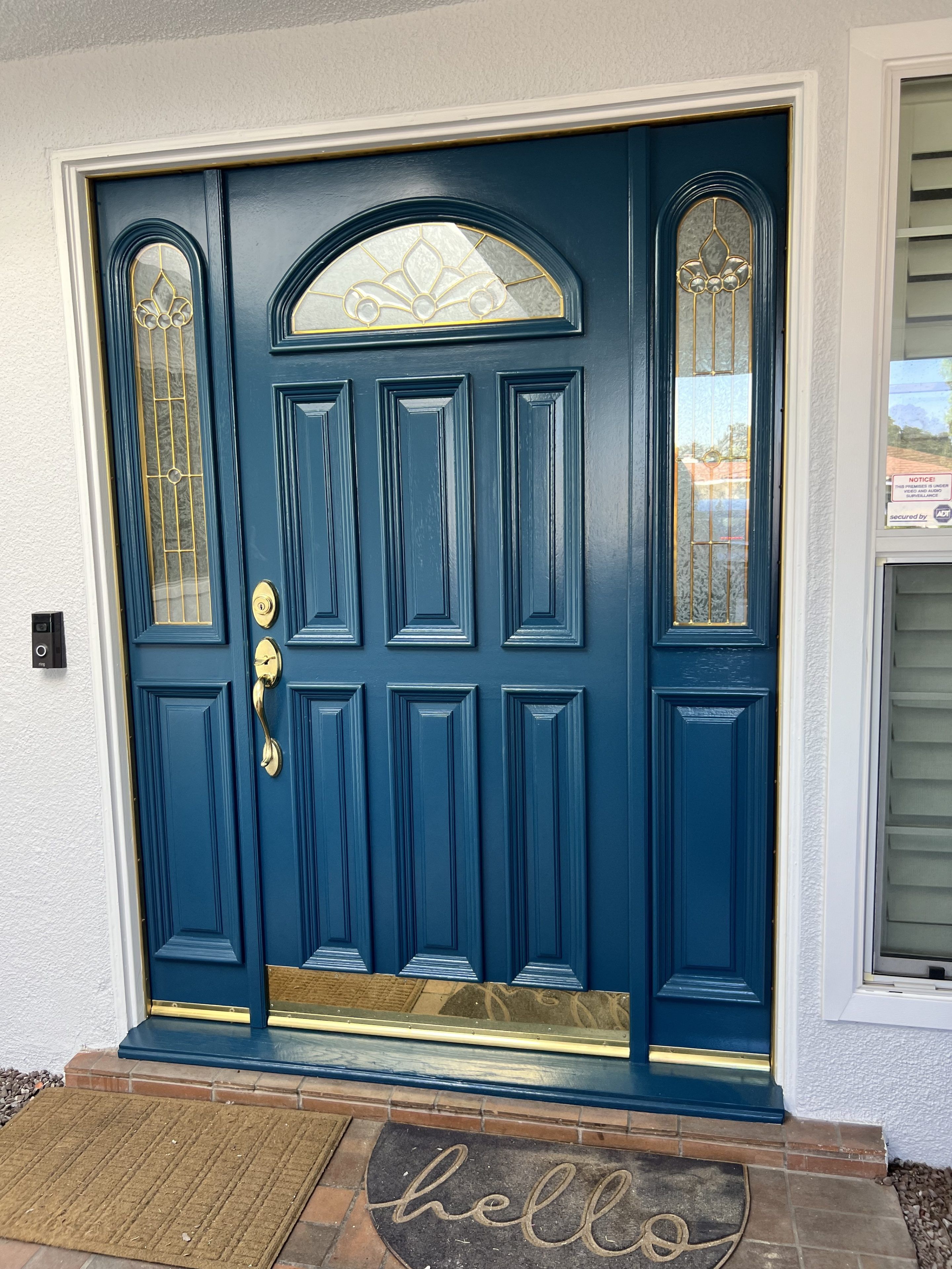 A teal front door with ornate glass panels and golden handle details is framed by a white wall. A doormat with "hello" is placed on the brick step.