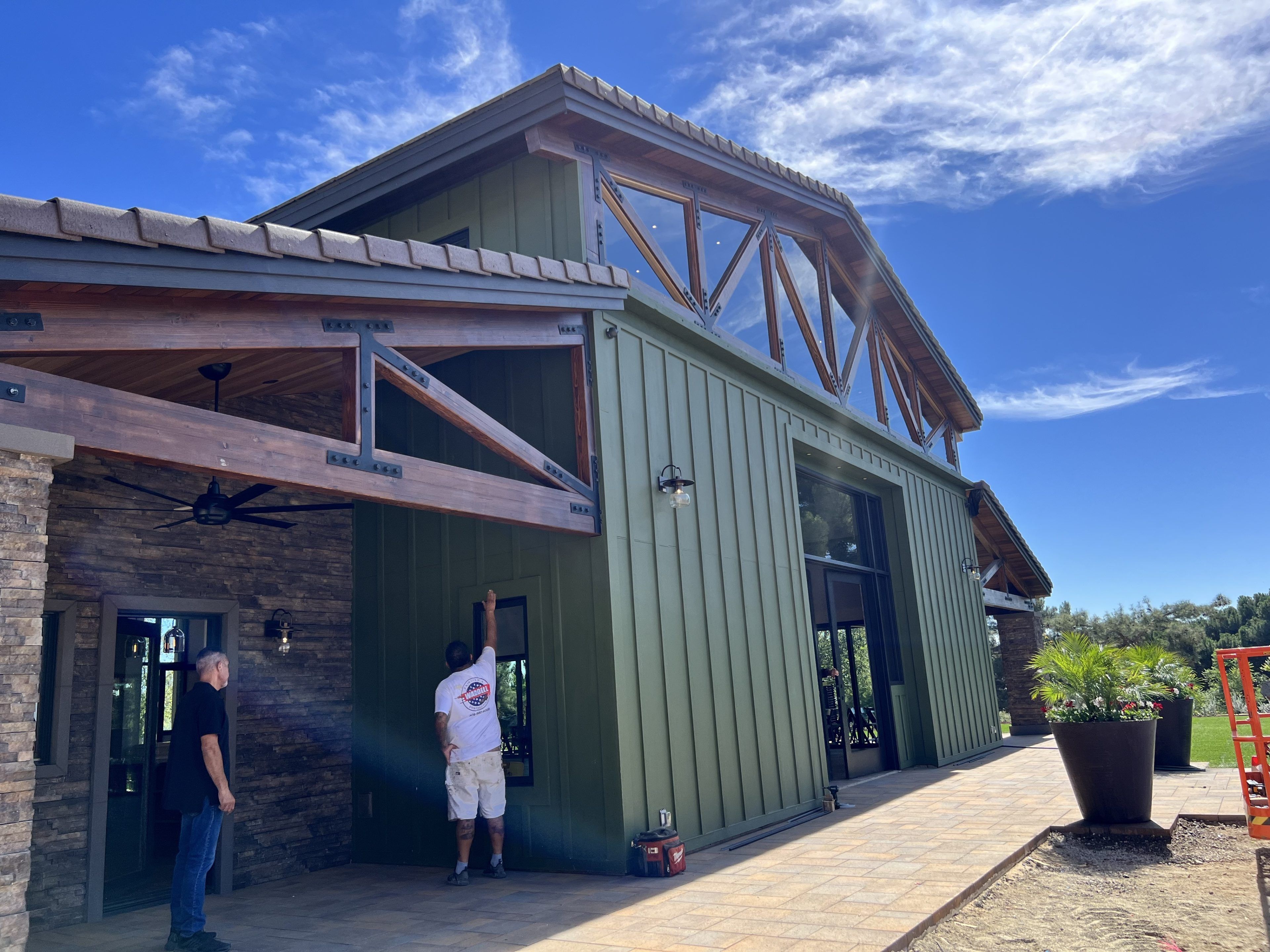 A modern green barn-style building with wooden beams and glass windows. Two people stand outside, observing the structure on a sunny day.