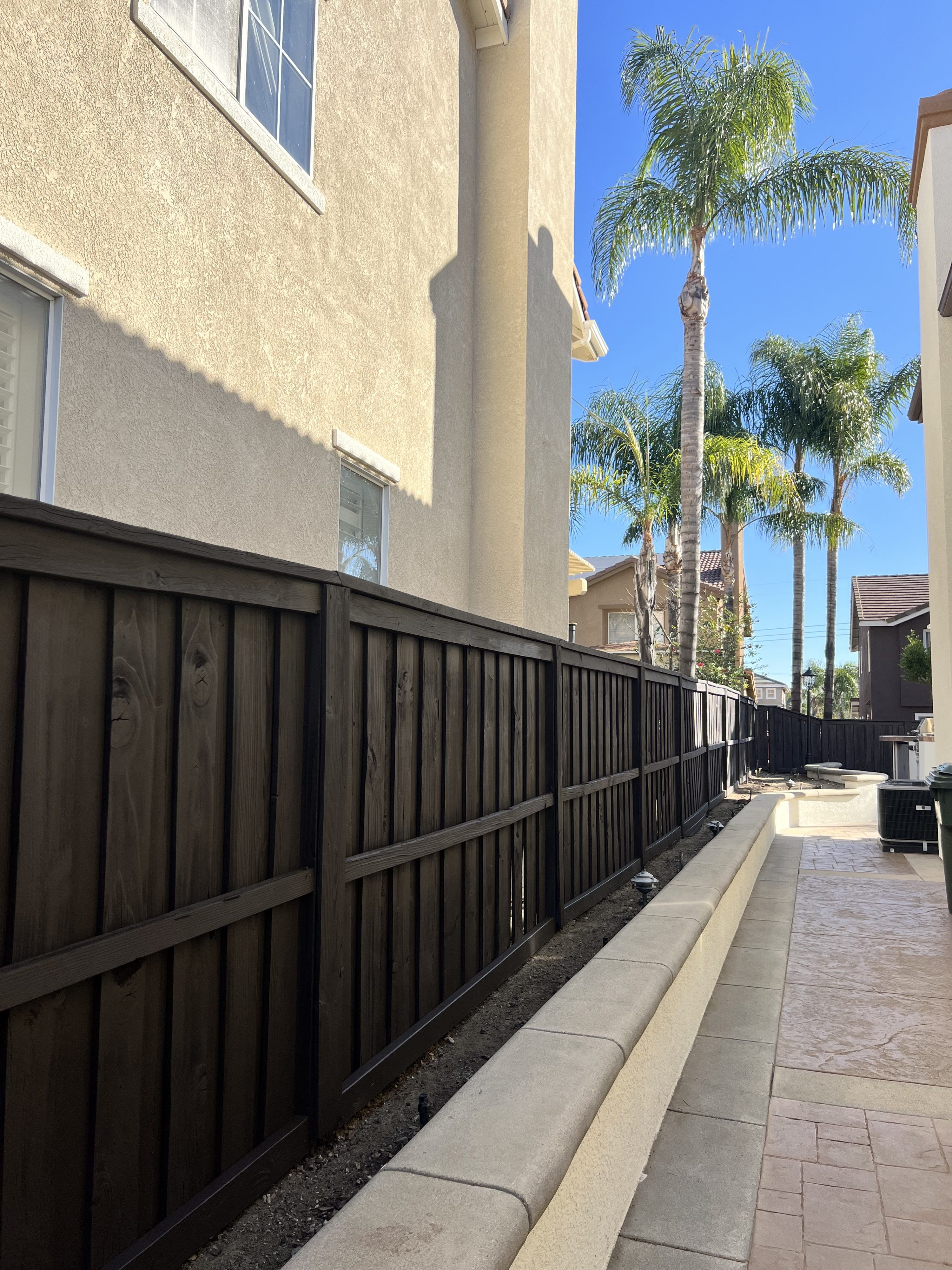 Narrow walkway between beige buildings with tall palm trees in the background. A dark wooden fence runs along the left side, creating a tranquil suburban feel.