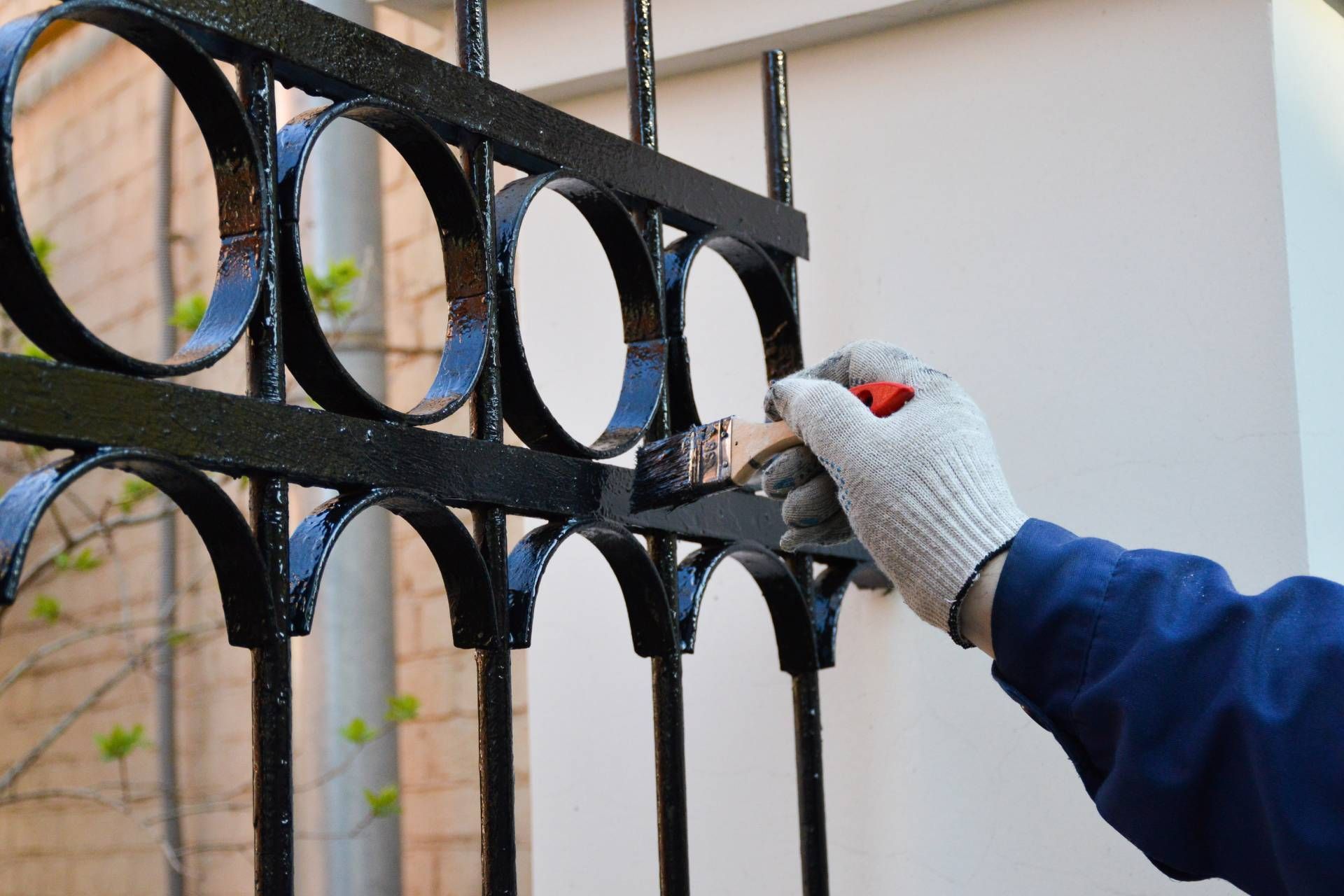 A gloved hand paints a black iron fence with a brush. The fence features circular patterns against a brick wall backdrop, conveying a sense of precision and care.