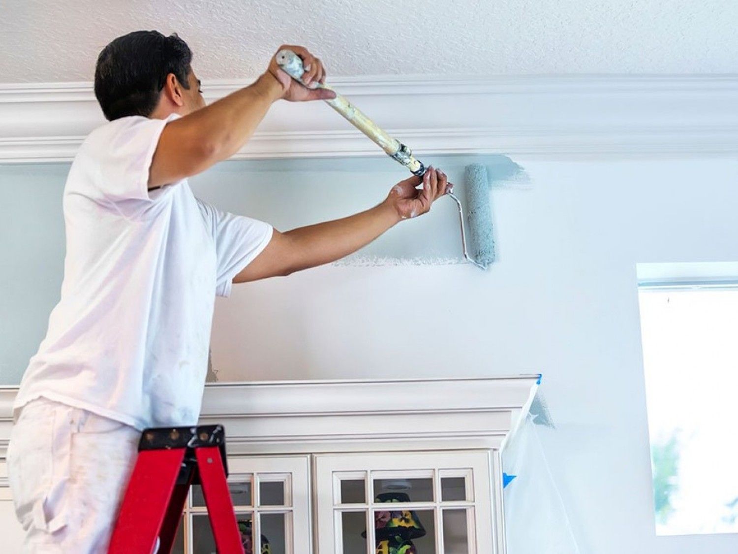 A person on a ladder paints a white wall with a blue roller brush near the ceiling. Light enters from a nearby window, creating a bright atmosphere.
