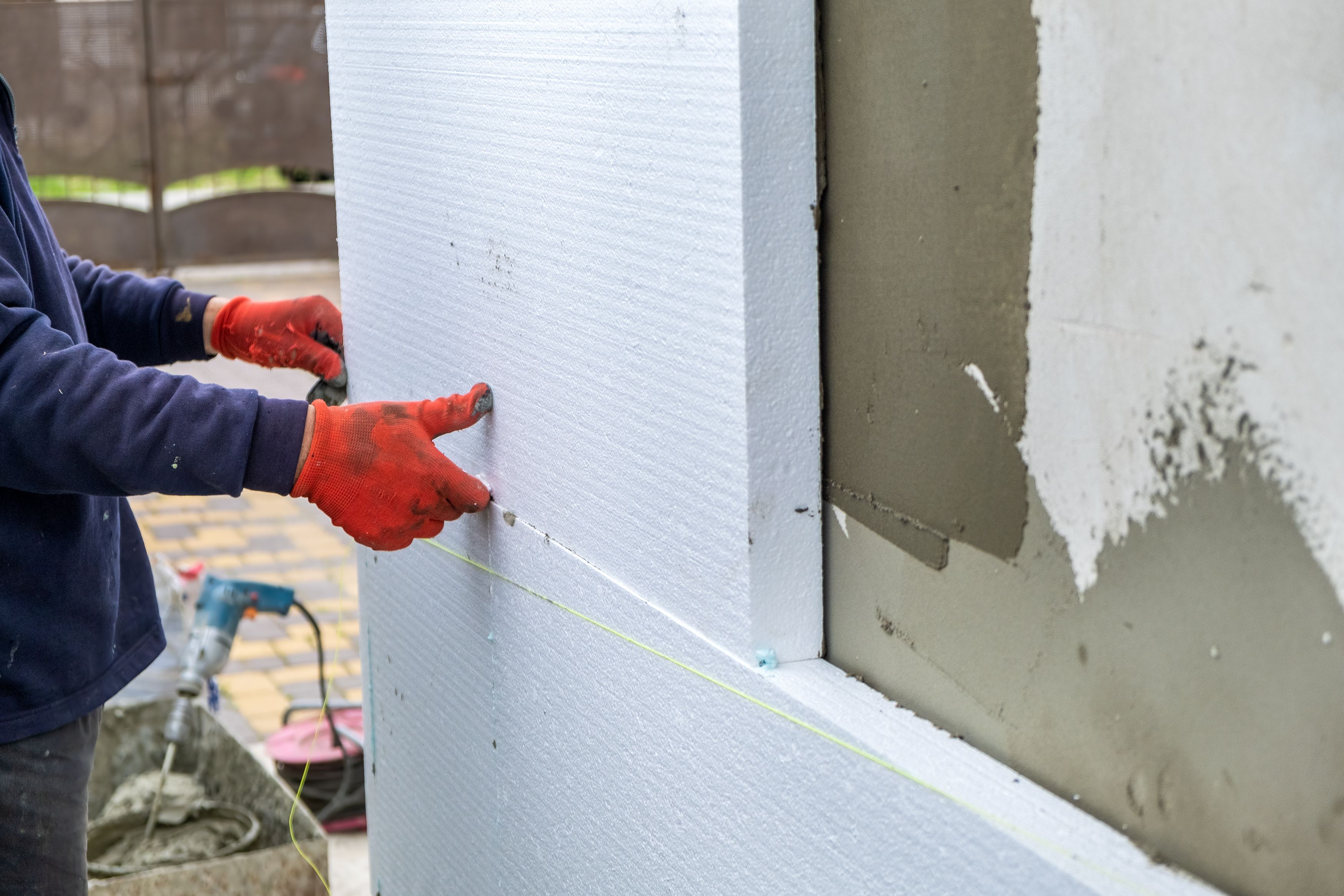 Worker wearing red gloves installs white foam insulation panels on an exterior wall, showing a close-up of hands aligning the panels with precision.
