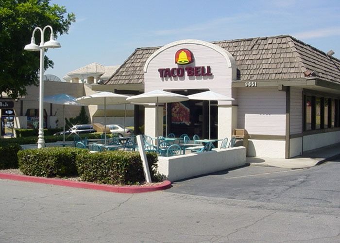 A Taco Bell with a vintage design features a red sign with a yellow bell on a wood-shingled roof. There is outdoor seating with tables and umbrellas.