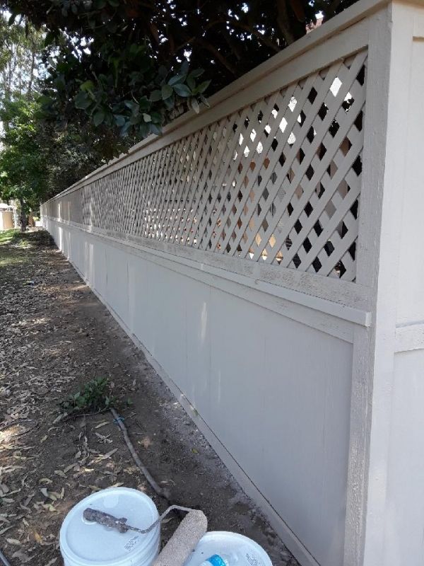 A long, freshly painted beige wooden fence stretches alongside a leaf-strewn path. Lattice design at the top. Paint cans and a roller rest nearby.
