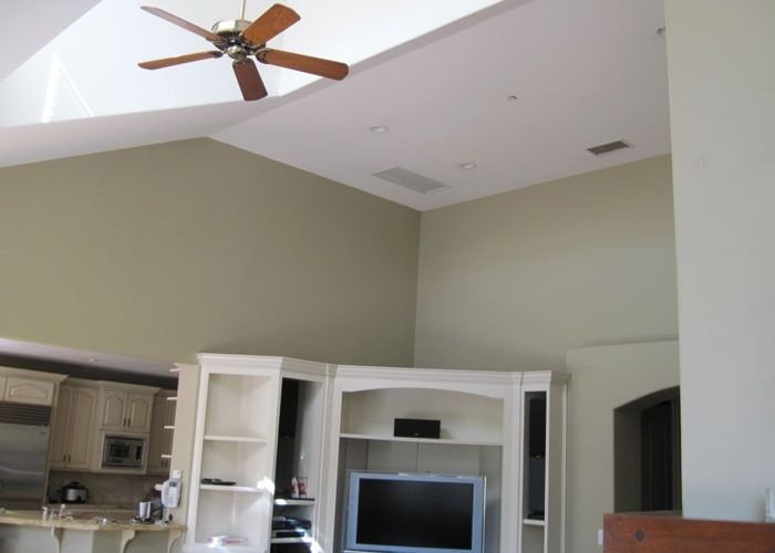 Spacious living room with high ceiling and a ceiling fan. Beige walls complement white cabinetry housing a flat-screen TV. Natural light fills the space.