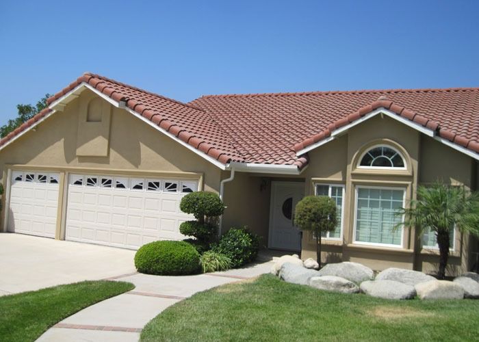 Single-story home with a red-tiled roof, cream stucco walls, and a two-car garage. Neat shrubs and rocks adorn the manicured lawn, evoking serene suburban life.