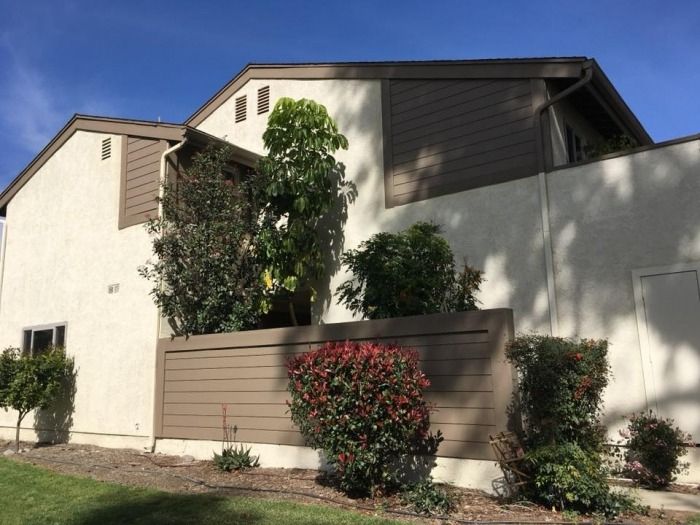 A beige, two-story house with a sloped roof and brown accents, surrounded by a tall wooden fence. Lush green bushes and a tree are in the sunlight.