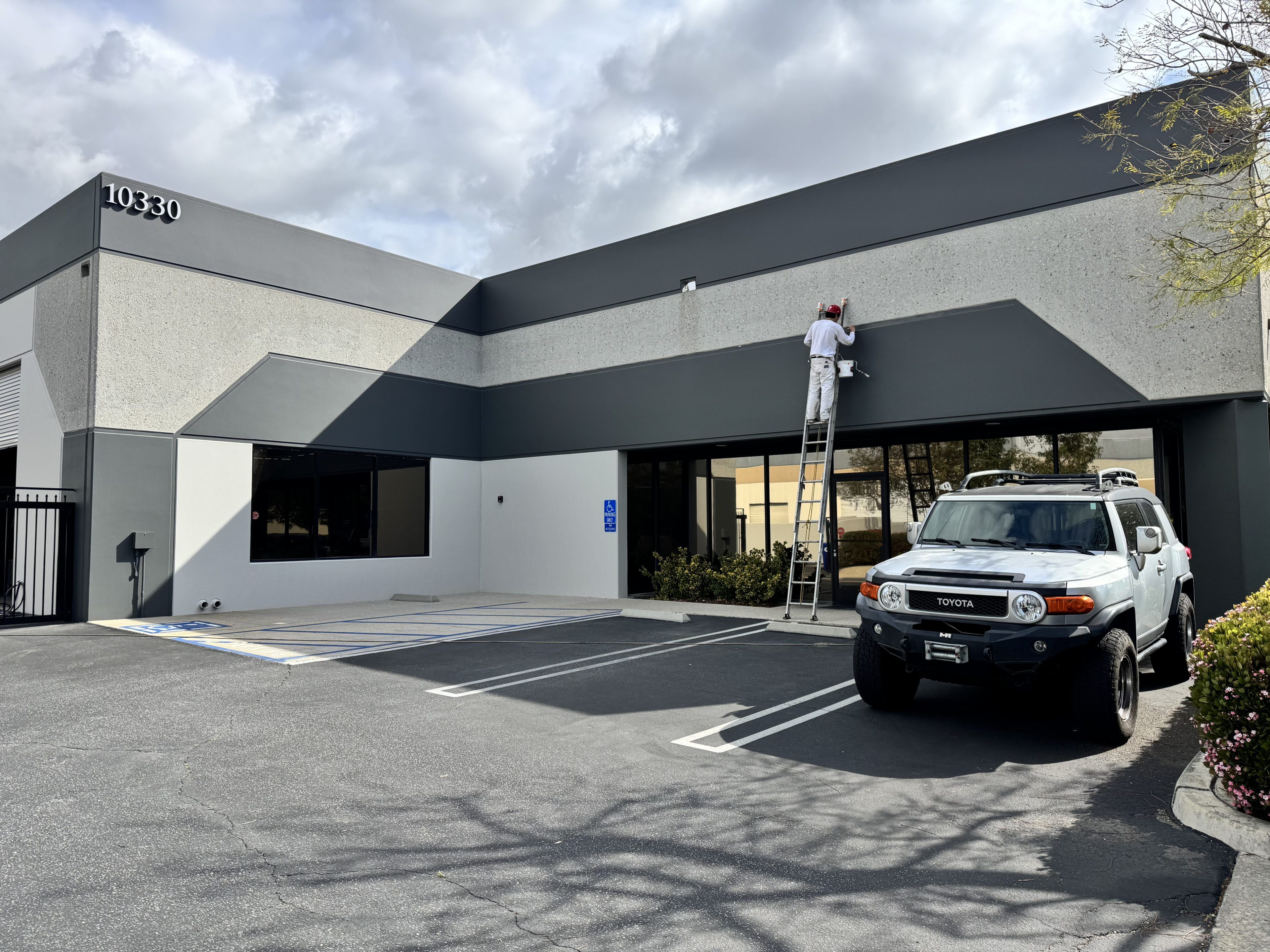 A person on a ladder paints a gray and white commercial building. An SUV is parked in front. The sky is partly cloudy, creating a calm atmosphere.